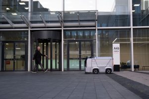 A modern office setting featuring a man and an autonomous delivery robot outside a glass-covered building