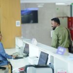A hotel receptionist works at a computer while a guest approaches the front desk, the kind of hotel system travel technology vendors sell.