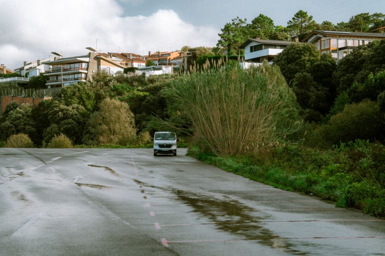 A white van sits on a quiet neighborhood road bordered by trees and homes